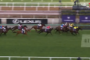 Group of horse racers sprinting on a green turf track; jockeys in colorful silks, banners and stands in the background.