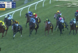 Group of colorful-jacketed jockeys ride closely packed horses along a turf racecourse, rail on left, with on-screen race timer and positions overlayed.
