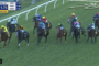 Group of colorful-jacketed jockeys ride closely packed horses along a turf racecourse, rail on left, with on-screen race timer and positions overlayed.