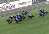 Pack of jockeys in colorful silks racing a grassy track, steady forward momentum past sponsor banners along the fence.