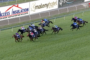 Pack of jockeys in colorful silks racing a grassy track, steady forward momentum past sponsor banners along the fence.
