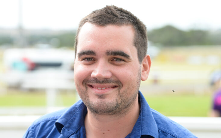 Close-up of a smiling man with short dark hair and stubble, wearing a blue shirt, outdoors on a sunny day