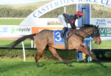 Jockey in red helmet rides a dark horse at full gallop on a race track with a large white arch in the background.