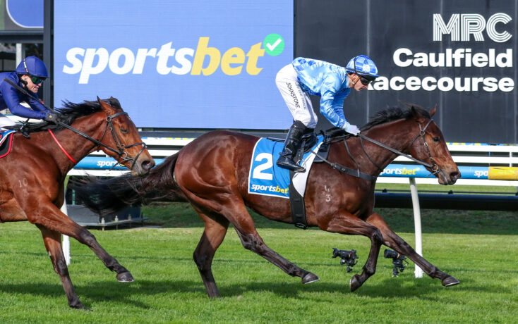 Two jockeys ride galloping horses during a race at Caulfield, with a large blue 'sportsbet' sign in the background.
