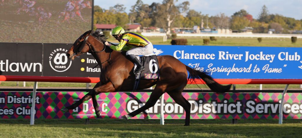 Jockey in neon green and purple silks rides a brown horse at full gallop along a racetrack with blue advertising banners in the background.
