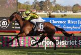 Jockey in neon green and purple silks rides a brown horse at full gallop along a racetrack with blue advertising banners in the background.