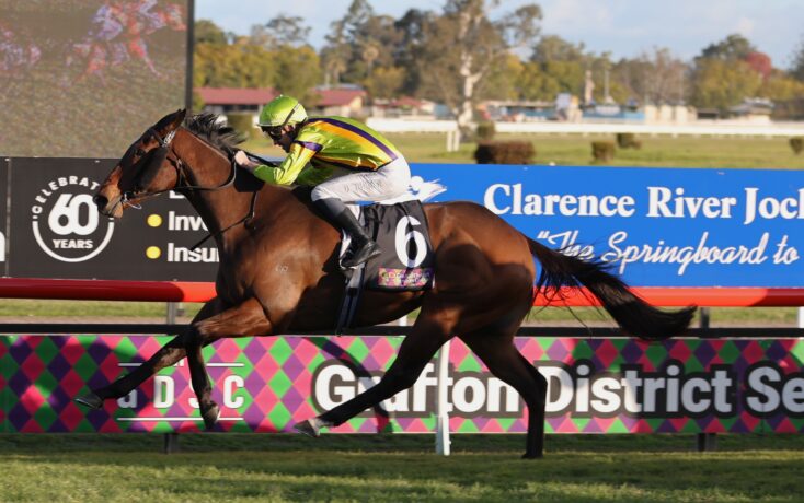 Jockey in neon green and purple silks rides a brown horse at full gallop along a racetrack with blue advertising banners in the background.