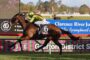 Jockey in neon green and purple silks rides a brown horse at full gallop along a racetrack with blue advertising banners in the background.