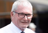 Smiling older man with gray hair and black-framed glasses, wearing a white shirt and tie outdoors.