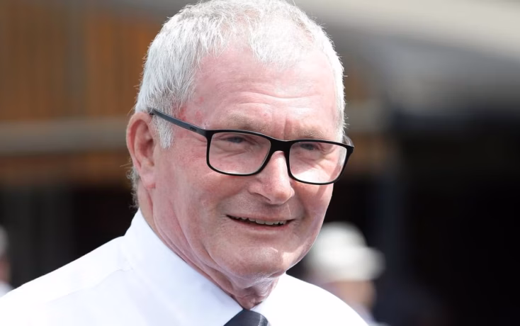 Smiling older man with gray hair and black-framed glasses, wearing a white shirt and tie outdoors.