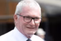 Smiling older man with gray hair and black-framed glasses, wearing a white shirt and tie outdoors.