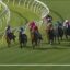 Group of jockeys on colorful silks racing around a curve on a grass track with white railings.