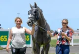 Two smiling women walk a gray horse on a lead at a sunny racetrack, with a betting sign in the background.