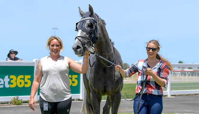Two smiling women walk a gray horse on a lead at a sunny racetrack, with a betting sign in the background.
