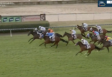 Group of racehorses with jockeys in colorful silks competing on a green turf track, near the rail; timer displayed in the top-right.