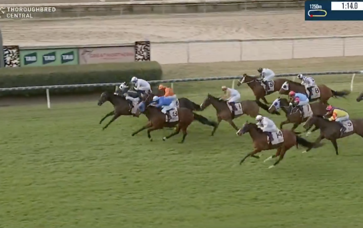 Group of racehorses with jockeys in colorful silks competing on a green turf track, near the rail; timer displayed in the top-right.