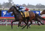 A jockey in blue-and-white silks rides a dark horse leading a race on a grassy track with colorful advertising hoardings in the background, closely followed by rivals.