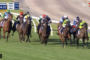 Several jockeys ride horses in a tight group on a grassy racecourse; clock and race data visible on screen.