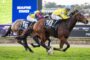 Two thoroughbred racehorses sprint neck-and-neck on a turf track, jockeys in bright silks (yellow/blue and red/blue) pushing to the finish at the Beaufine Stakes display in the background.