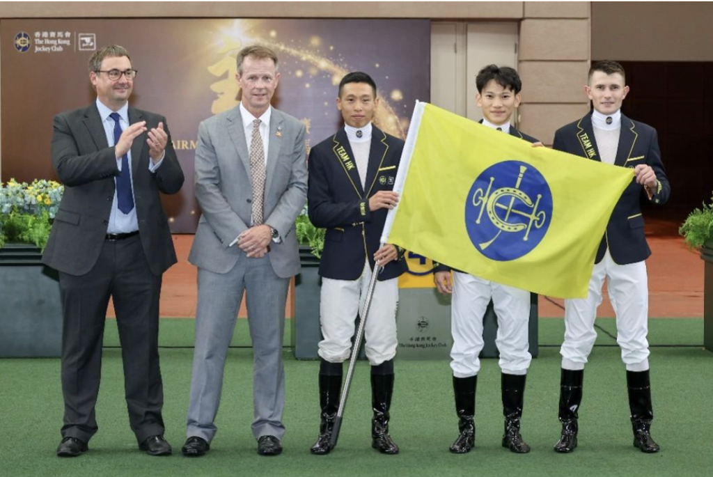 Five people posing at a ceremony: two suited men on the left applauding, two jockeys in navy jackets and white breeches holding a yellow flag with a blue emblem, with a sponsor backdrop.