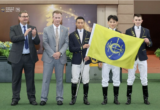 Five people posing at a ceremony: two suited men on the left applauding, two jockeys in navy jackets and white breeches holding a yellow flag with a blue emblem, with a sponsor backdrop.