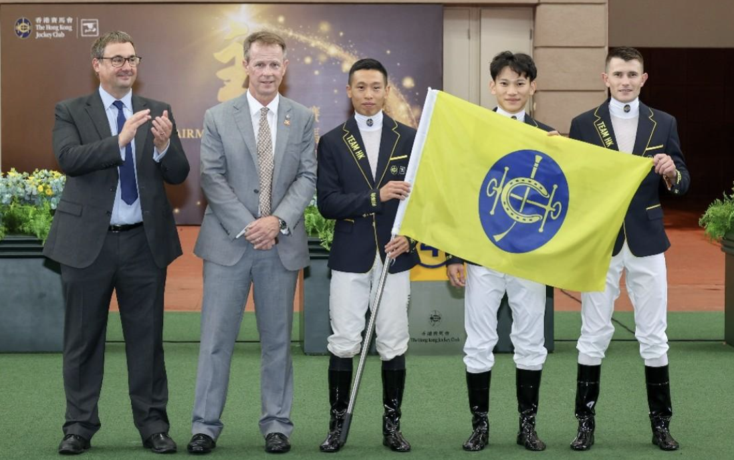 Five people posing at a ceremony: two suited men on the left applauding, two jockeys in navy jackets and white breeches holding a yellow flag with a blue emblem, with a sponsor backdrop.