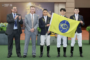 Five people posing at a ceremony: two suited men on the left applauding, two jockeys in navy jackets and white breeches holding a yellow flag with a blue emblem, with a sponsor backdrop.