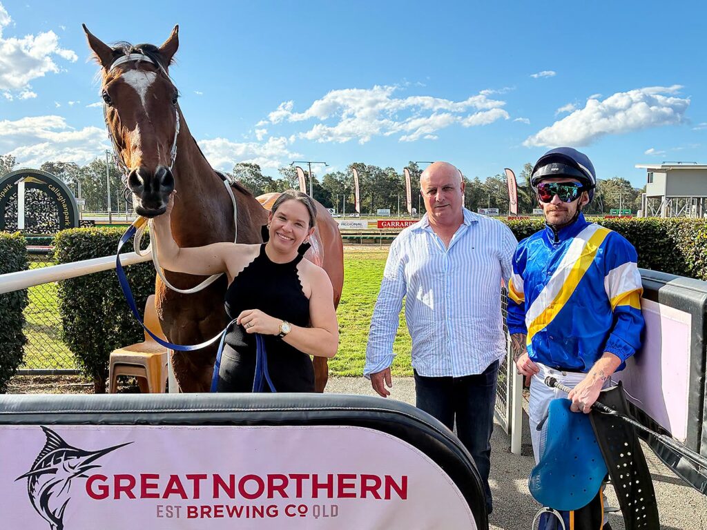 John Dann Illegal Justice | Just Horse Racing Woman in a black sleeveless top holds the reins of a brown horse beside a man in a striped shirt and a jockey in blue‑yellow silks at a sunny racecourse.