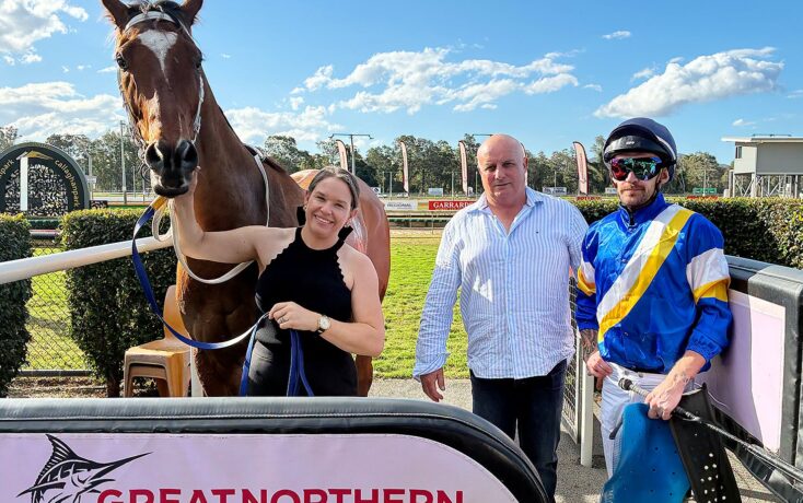 Woman in a black sleeveless top holds the reins of a brown horse beside a man in a striped shirt and a jockey in blue‑yellow silks at a sunny racecourse.