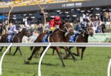 Jockeys and horses racing on a green turf track with a crowd in the background and 'J.R. Fleming Stand' signage visible in the stands.