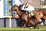 Two racehorses with jockeys lean forward as they sprint on a grassy track near a blue barrier with letters behind them.