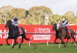 Two jockeys race on horseback toward a red advertising barrier during a horse race; black horse leads on left, chestnut horse on right.