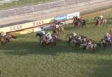 Horses and jockeys race around a bend on a grassy turf track with rail and advertising boards along the inside fence