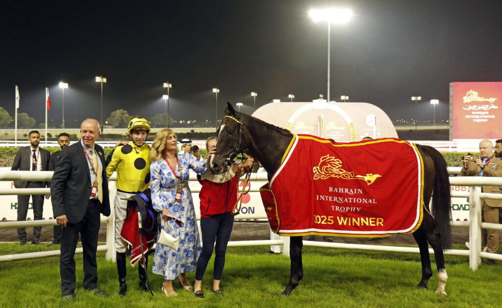 ROYAL CHAMPION | Just Horse Racing Nighttime paddock scene with a dark horse wearing a red blanket that reads Bahrain International Trophy; people in suits and a woman in a blue patterned dress surround the horse, smiling and photographing."