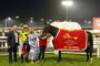 Nighttime paddock scene with a dark horse wearing a red blanket that reads Bahrain International Trophy; people in suits and a woman in a blue patterned dress surround the horse, smiling and photographing."