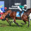 Two jockeys in blue and green silks race their horses on a grass track at Caulfield Racecourse, with a large sponsor banner in the background.