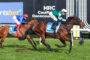 Two jockeys in blue and green silks race their horses on a grass track at Caulfield Racecourse, with a large sponsor banner in the background.