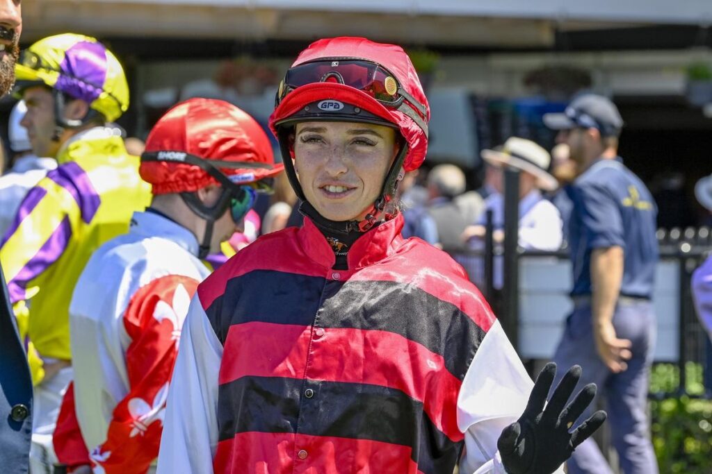 Siena Grima | Just Horse Racing Jockey in red and black striped silks and helmet waves to camera at a racecourse surface setting.