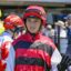 Jockey in red and black striped silks and helmet waves to camera at a racecourse surface setting.
