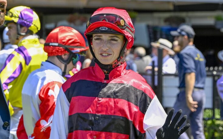 Jockey in red and black striped silks and helmet waves to camera at a racecourse surface setting.