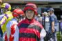 Jockey in red and black striped silks and helmet waves to camera at a racecourse surface setting.