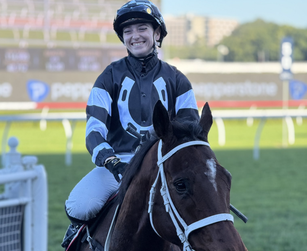 Jockey in blue-and-black racing silks smiles while riding a dark brown horse on a race track.