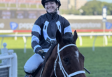 Jockey in blue-and-black racing silks smiles while riding a dark brown horse on a race track.