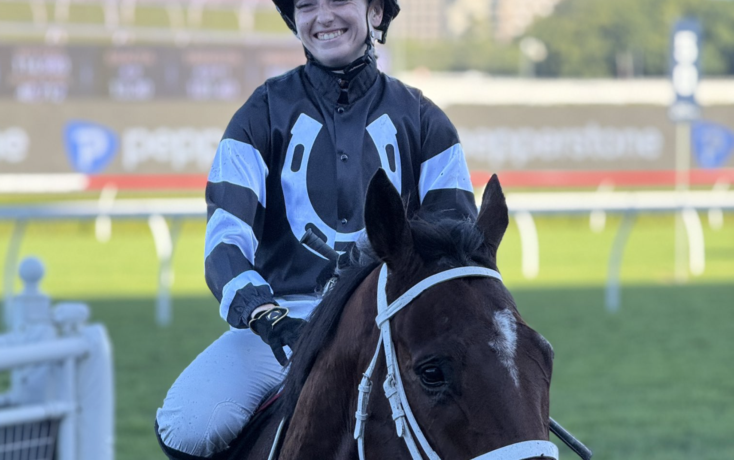Jockey in blue-and-black racing silks smiles while riding a dark brown horse on a race track.