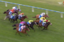 Horses race on a green turf track with jockeys in colorful silks, a tight pack sprinting toward the finish line.