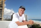 Smiling man in a white polo leaning on a balcony railing with a beach and ocean in the background.