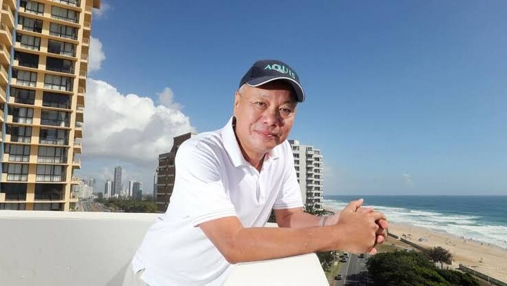 Smiling man in a white polo leaning on a balcony railing with a beach and ocean in the background.