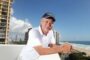 Smiling man in a white polo leaning on a balcony railing with a beach and ocean in the background.