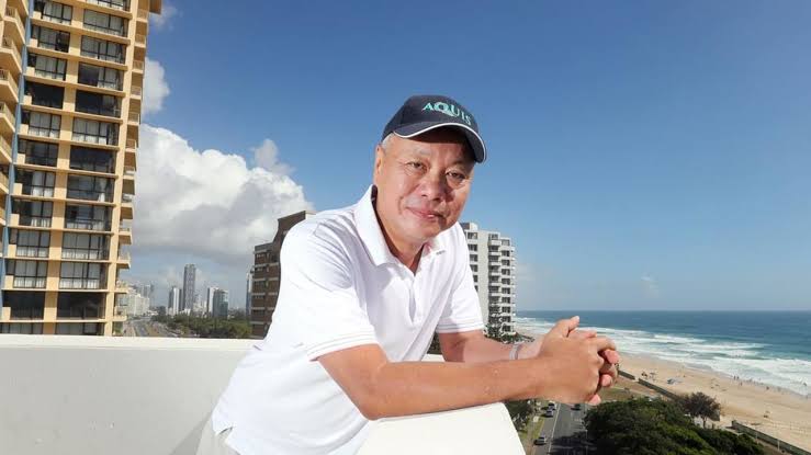 Smiling man in a white polo leaning on a balcony railing with a beach and ocean in the background.