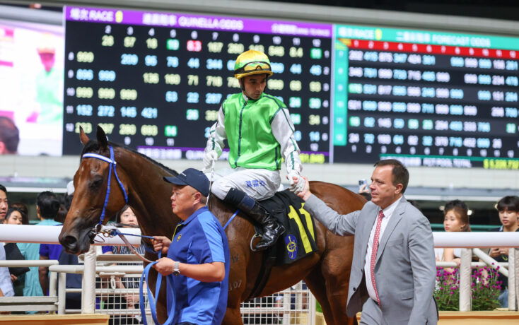 Jockey in bright green vest wearing a helmet on a brown horse being guided by handlers near a crowded railing with a large scoreboard in the background